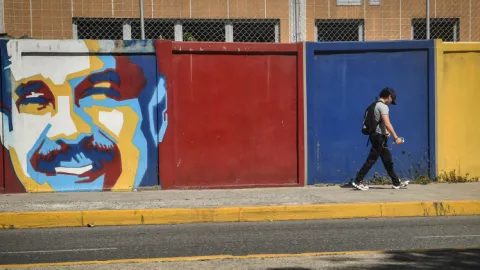 Colorful mural of a man’s face painted on a wall beside a red and blue gate, with a person walking past on the sidewalk. 